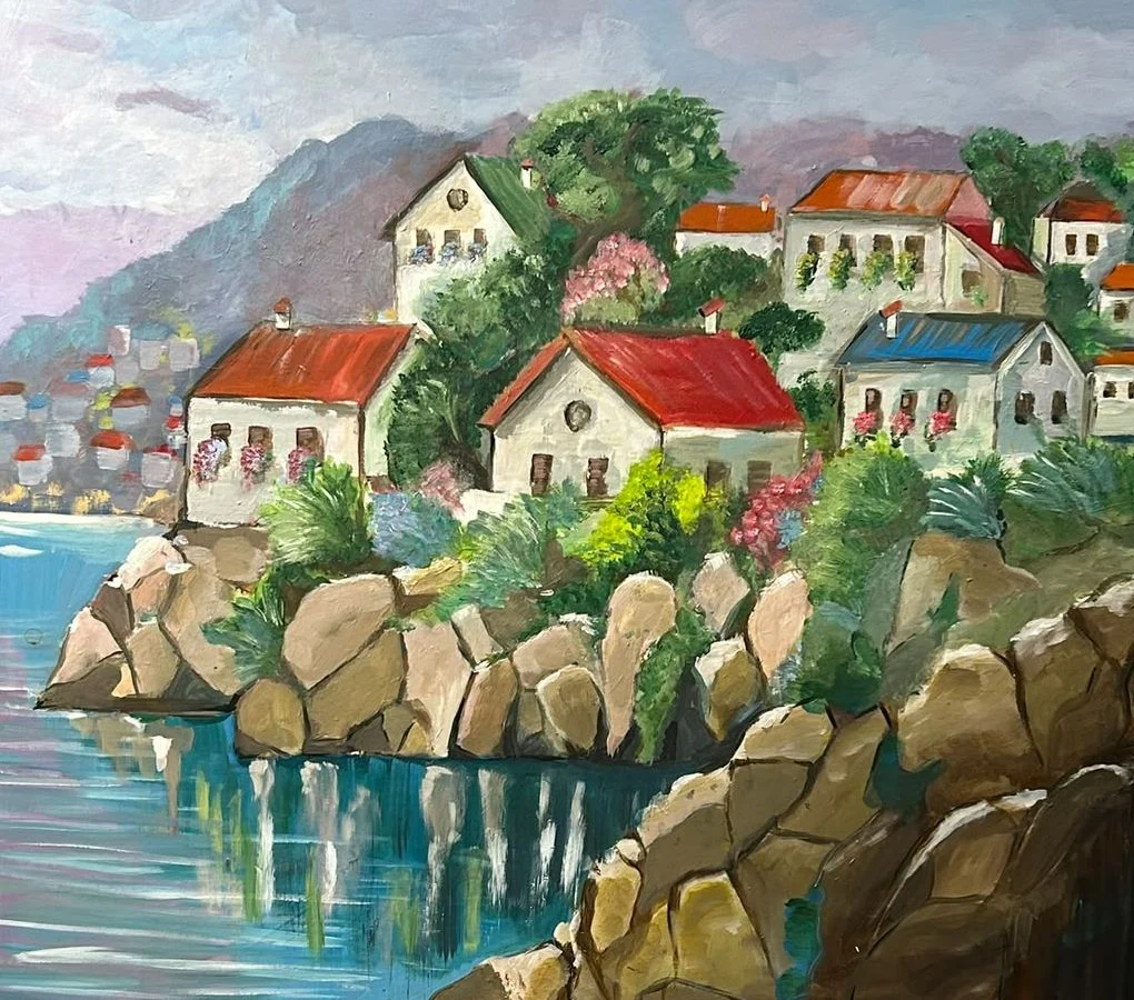 White houses with terracotta and blue roofs sit among trees above a rocky shoreline, reflected in turquoise coastal water.